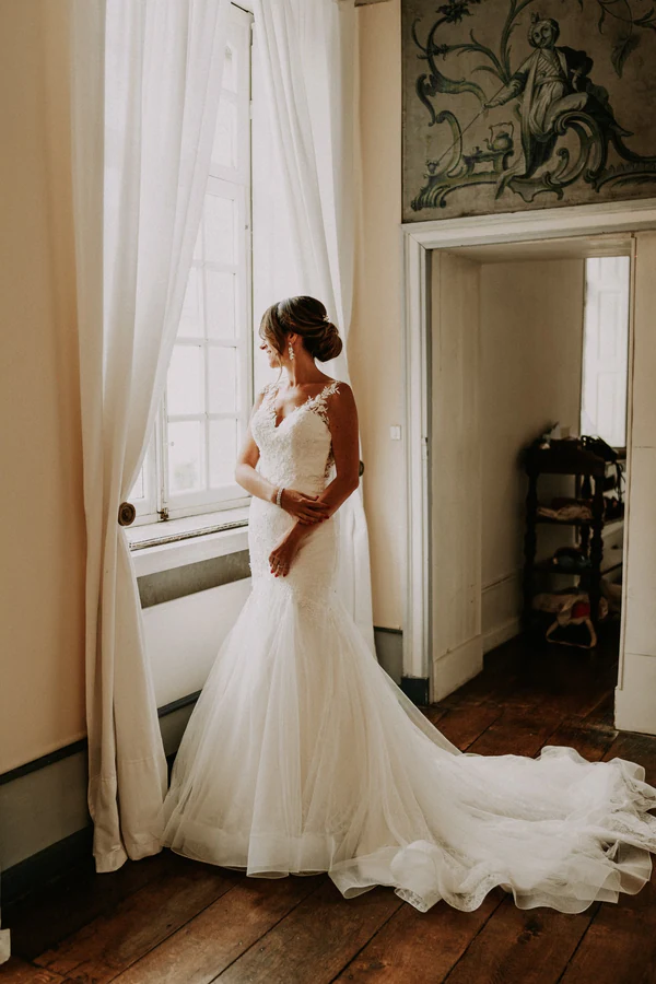 La mariée et sa robe à traîne devant une grande fenêtre, mariage au Château de Deulin