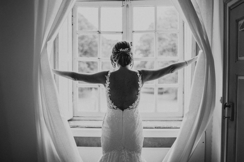 La mariée face à la fenêtre, bras ouverts, en noir et blanc, mariage au Château de Deulin
