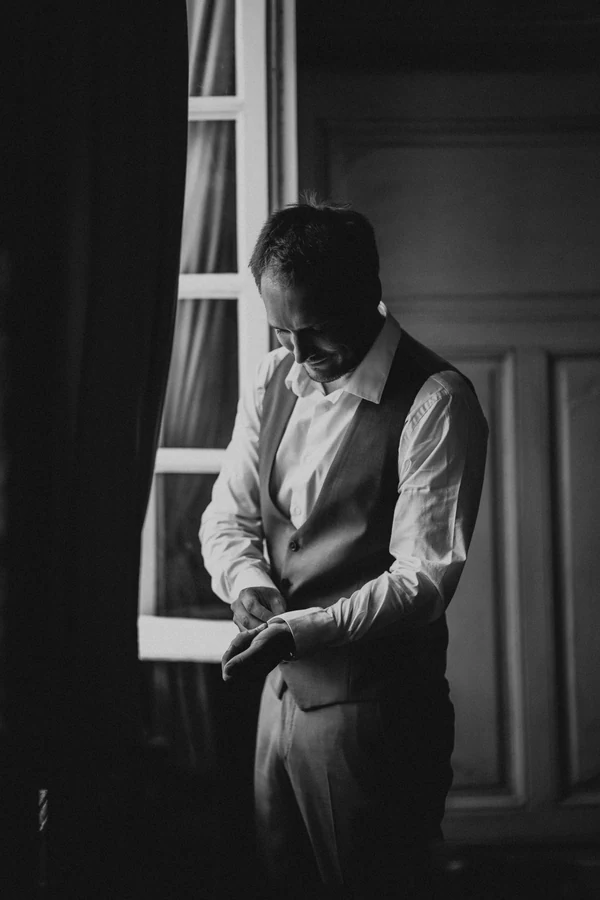 Le marié ajuste sa manchette près de la fenêtre, noir et blanc, mariage à Deulin
