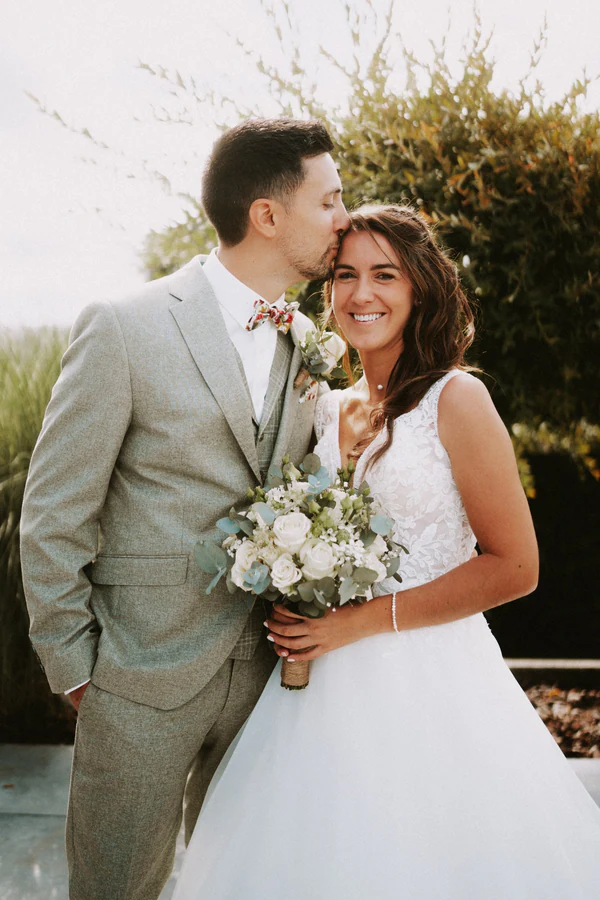 Le marié embrasse la mariée sur le front, bouquet blanc à la main, mariage au Primordia