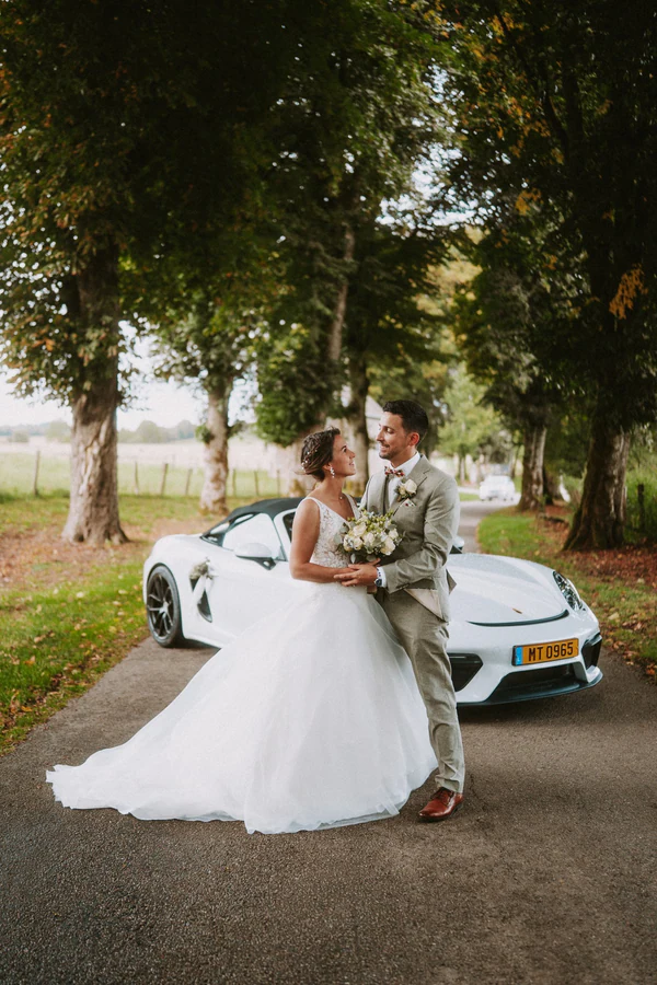 Les mariés devant un cabriolet blanc sur une allée bordée d'arbres, mariage au Primordia