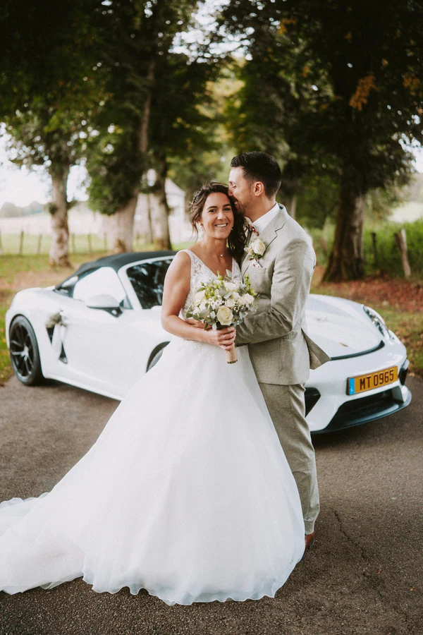 Les mariés enlacés près du cabriolet blanc, allée d'arbres, mariage au Primordia
