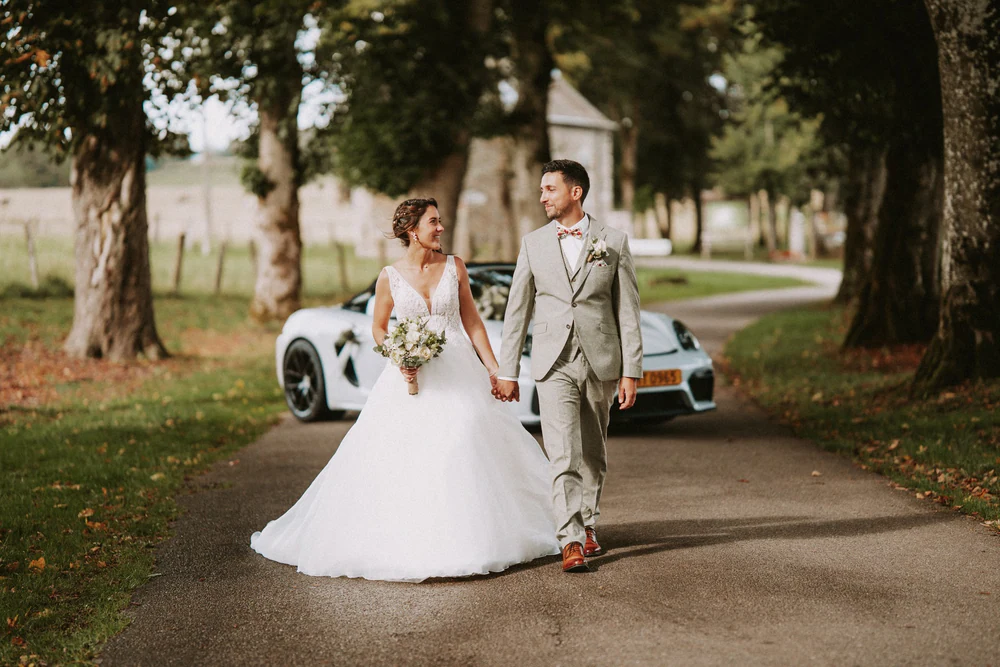 Les mariés main dans la main sur l'allée arborée, avec leur cabriolet blanc, mariage au Primordia