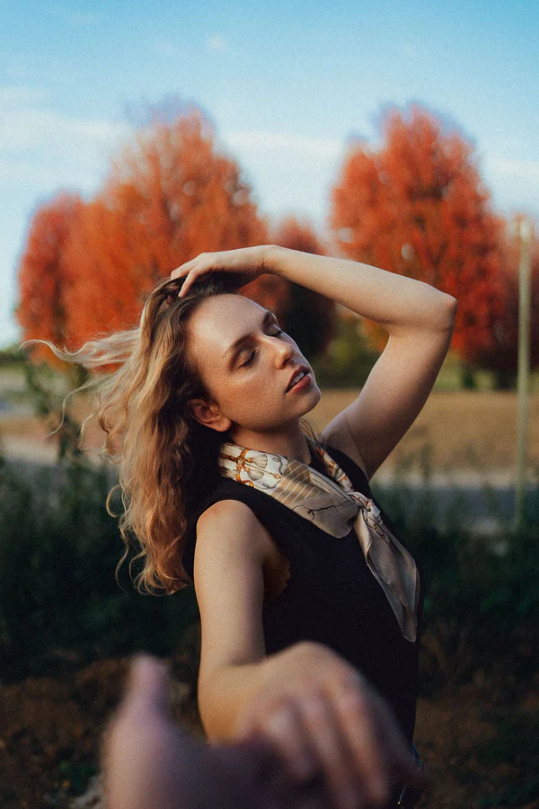 Portrait de Chloé les yeux fermés, main dans les cheveux au vent, foulard et arbres roux d'automne, par Gautier Collet