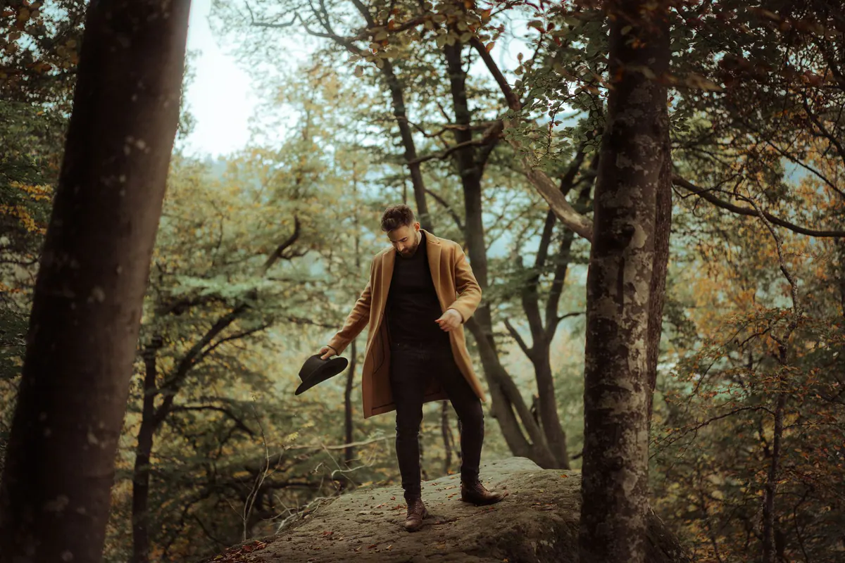 Gautier Collet en manteau camel, un chapeau à la main, sur un rocher en forêt à l'automne
