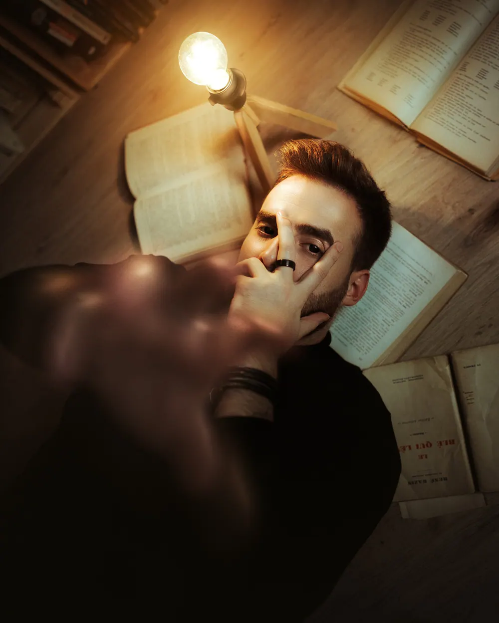 Gautier Collet allongé parmi de vieux livres, une ampoule allumée à côté de lui, la main devant le visage