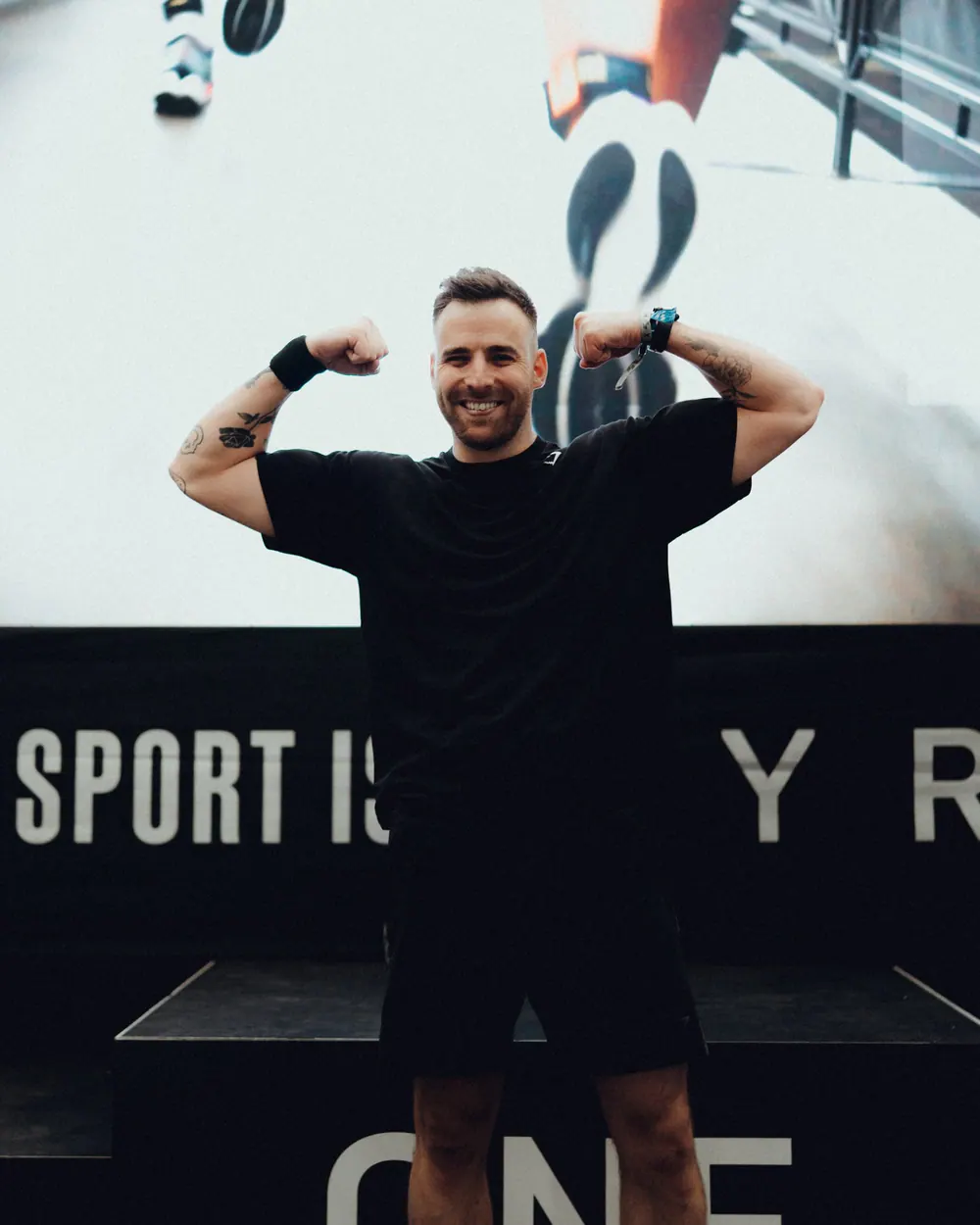 Gautier Collet souriant, les bras levés pour montrer ses biceps, devant un grand écran